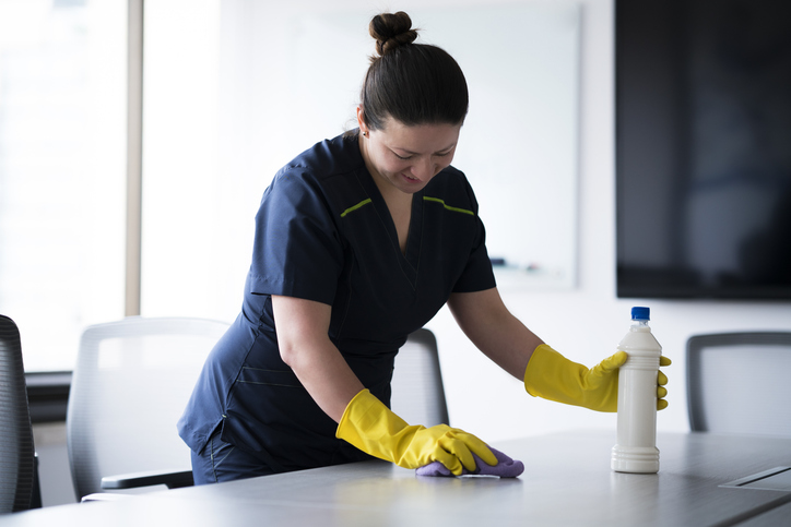 Woman doing the cleaning of a desk in an office, with her cleaning uniform, smiling doing this action